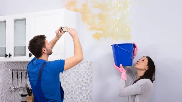 Couple photographing a ceiling leak while holding a bucket, documenting damage for a claim