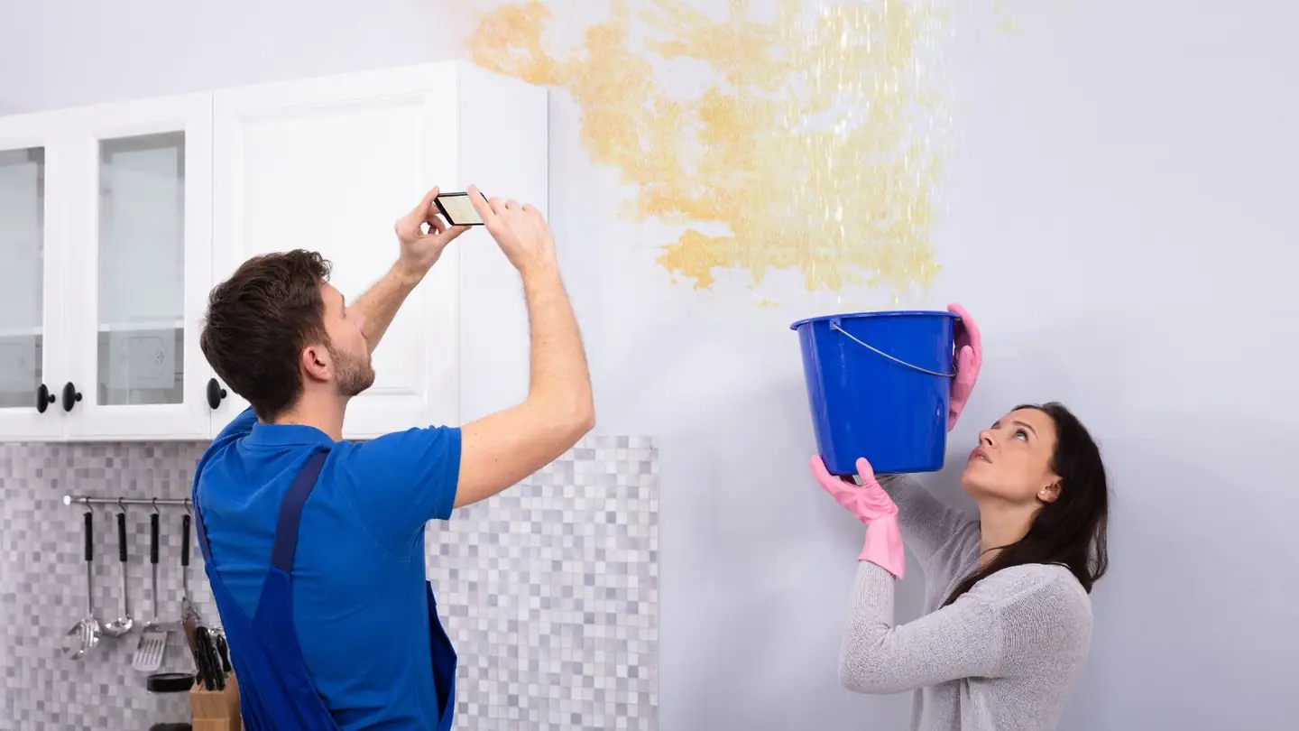 Couple photographing a ceiling leak while holding a bucket, documenting damage for a claim